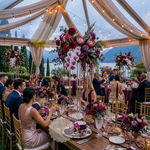 Load image into Gallery viewer, Dining table set for a formal event with floral arrangements and guests seated, under a tented canopy.

