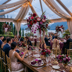Load image into Gallery viewer, Dining table set for a formal event with floral arrangements and guests seated outdoors.
